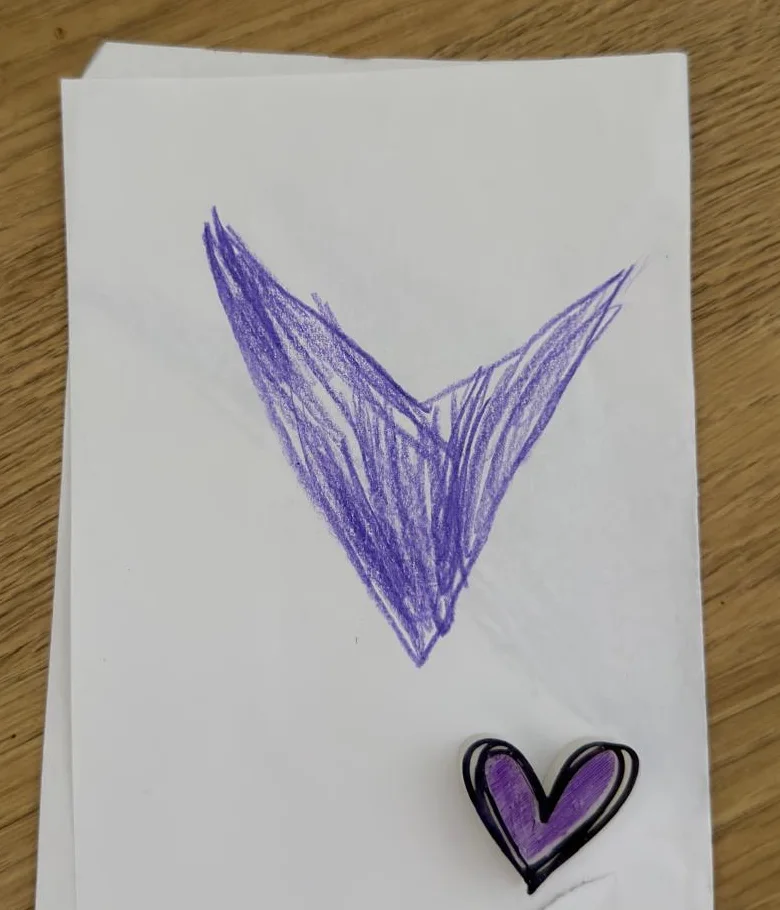 Un cuore viola disegnato a pastello su un foglio, e accanto il piccolo cuore stampato e dipinto con gli stessi colori.